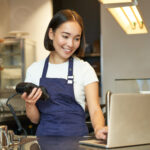 Smiling asian barista, girl with card terminal, payment machine and laptop, standing in cafe, processing payment for coffee order